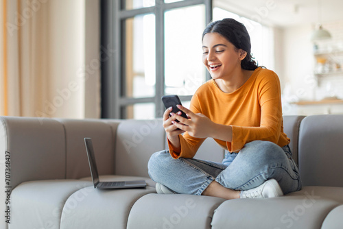 Happy indian woman using smartphone, sitting on sofa with laptop beside her