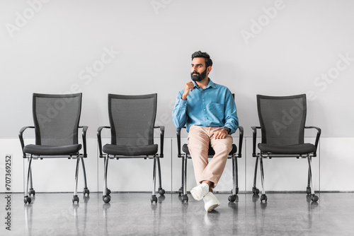 Pensive indian man in office with empty chairs, contemplating photo