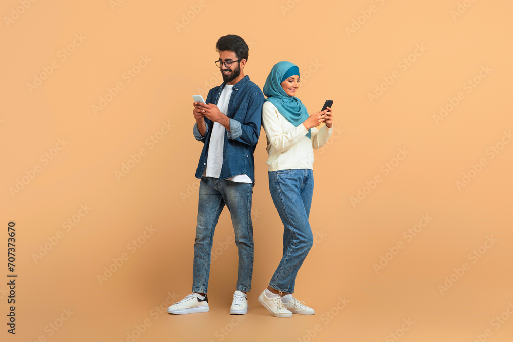 Modern arab muslim couple with their smartphones standing over beige background Modern arab muslim couple with their smartphones standing over beige background