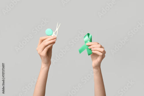 Female hands with green ribbon, container for contact lenses and tweezers on grey background. Glaucoma awareness concept