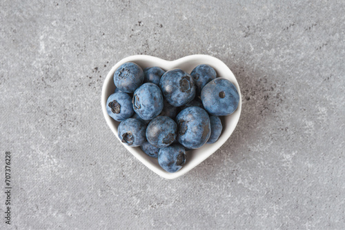 Fresh blueberries in a small heart-shaped bowl on gray concrete background. Organic berries, healthy food, wild berries. Top view, flat lay