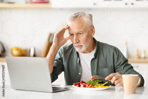 Concentrated senior man multitasking  eating lunch and working on laptop in kitchen