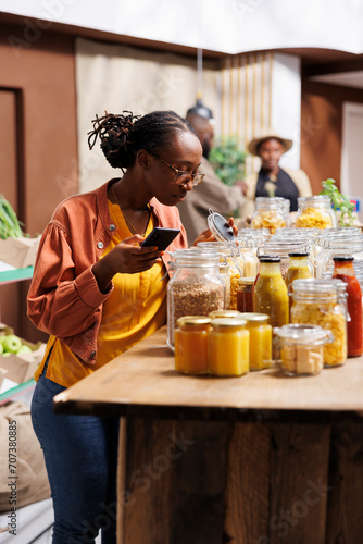African American female customer compares prices, using her smartphone in a modern supermarket. Black lady inspecting things in glass containers at a bio-food store while holding a mobile device.