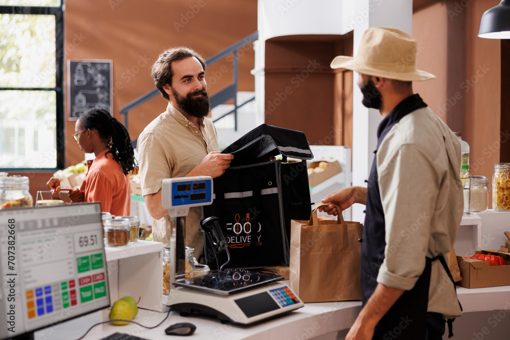 Caucasian male courier arrives at checkout counter ready to receive packaged grocery order from cashier. Food delivery man with backpack retrieves takeaway from vendor at local market. - obrazy, fototapety, plakaty 