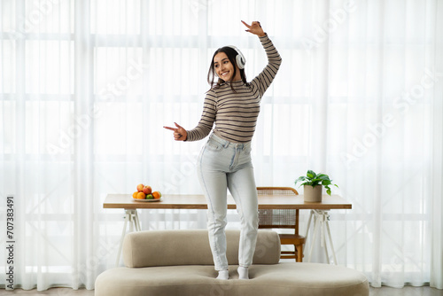Joyful young woman with headphones dancing on a beige couch, throwing her arms up