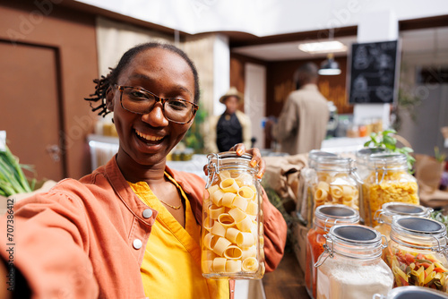 POV of a black woman wearing glasses, holding a glass container filled with pasta in a bio-food supermarket. Online marketing, organic items, vlogging, digital camera, customer filming.