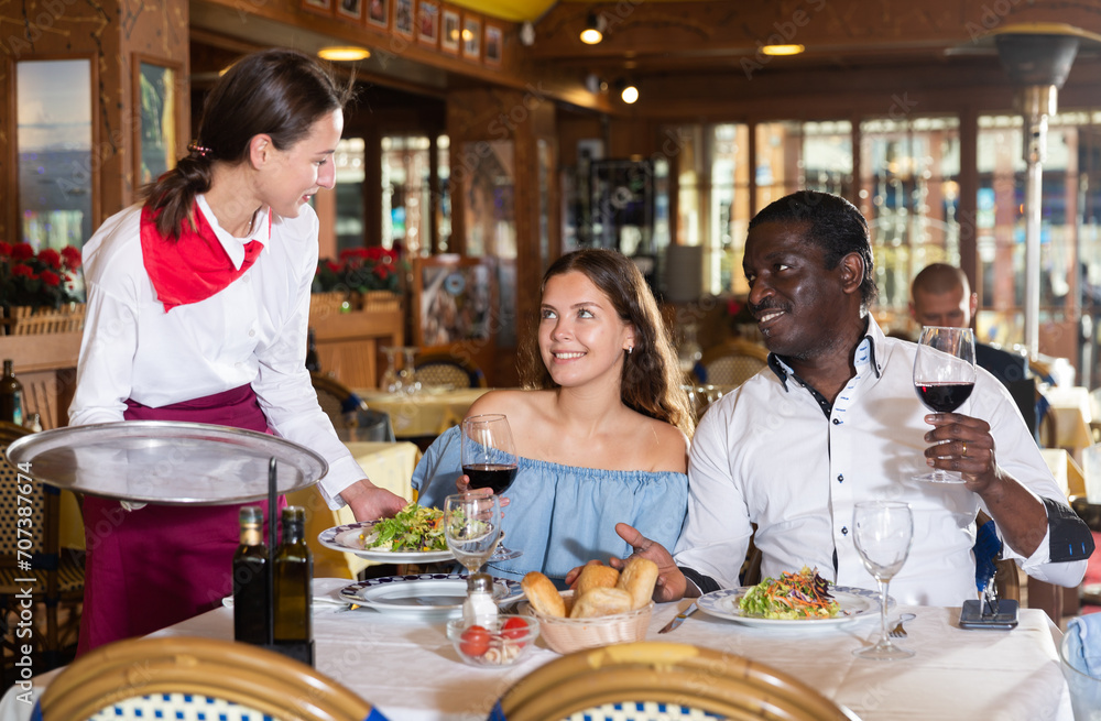Hospitable young beautiful waitress, bringing dishes to couple of guests at restaurant