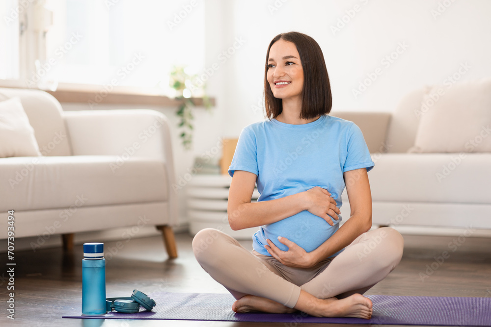 Pregnant lady exercising on floor and hugging her tummy indoors Pregnant lady exercising on floor and hugging her tummy indoors