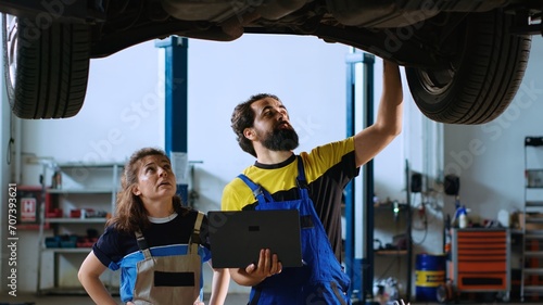 Certified serviceman in garage workspace standing with customer underneath suspended car, looking together for replacement parts. Expert helps woman fix vehicle, using laptop to find components online