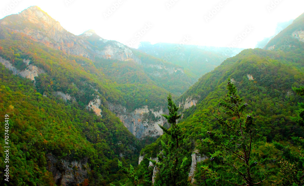 Fresh view at the impressive Sibillini Mountains in the gorge of Eremo di Soffiano near Sarnano with their rocky appearance sweetened by the rich vegetation, haze in background, trees in foreground Fresh view at the impressive Sibillini Mountains in the gorge of Eremo di Soffiano near Sarnano with their rocky appearance sweetened by the rich vegetation, haze in background, trees in foreground
