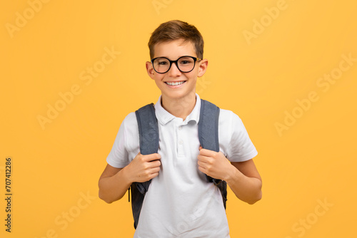 Cheerful schoolboy wearing glasses and backpack standing confidently on yellow studio background
