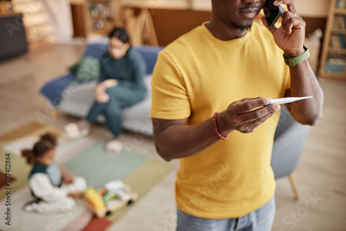 Cropped shot of African American father calling doctor using smartphone while holding digital thermometer standing in living room  mother and child in blurred background