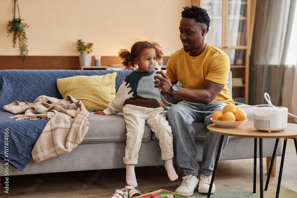 Full shot of smiling African American dad helping ill little daughter with inhalation through nebulizer while sitting on big sofa at home