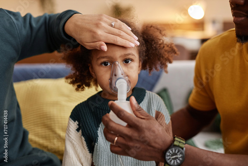 Portrait of little African American girl looking at camera while unrecognizable mother placing hand on her forehead and dad putting nebulizer mask on face photo