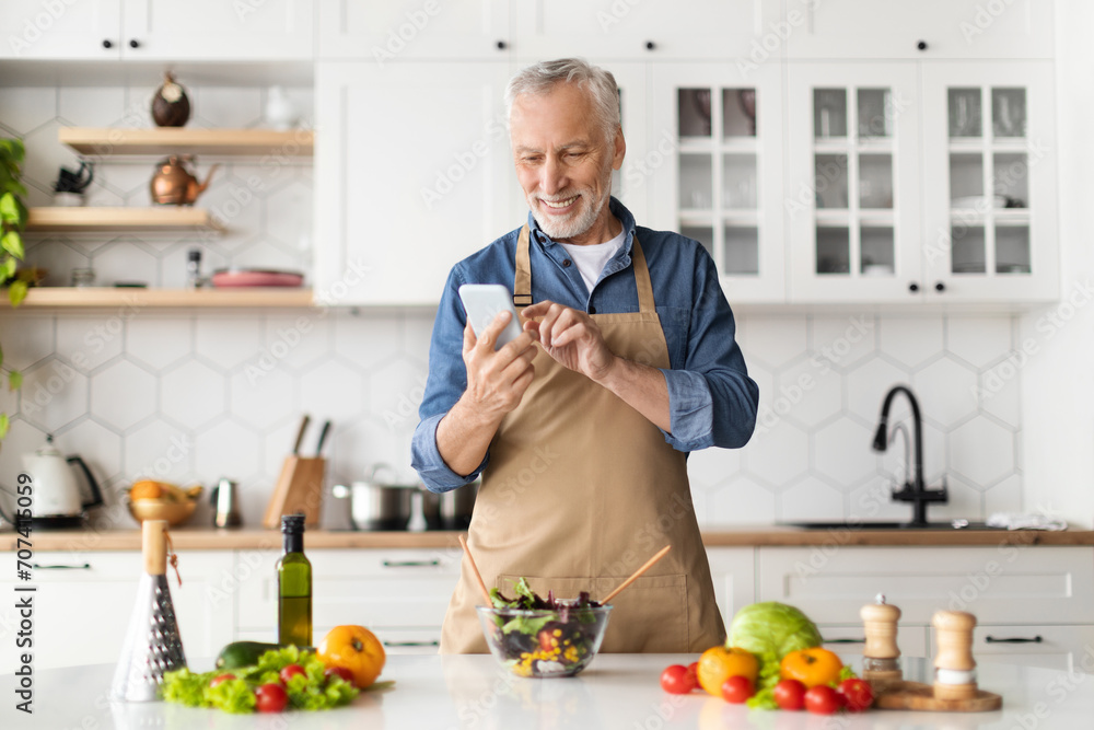 Cheerful senior man in apron using smartphone while cooking in kitchen Cheerful senior man in apron using smartphone while cooking in kitchen