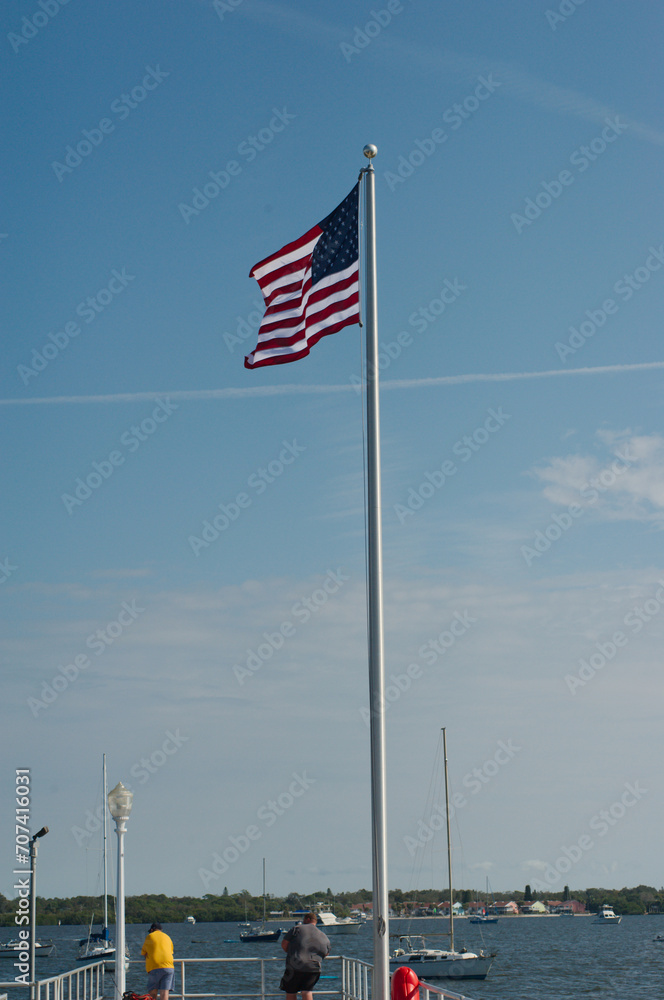 American Flag against blue sky with white clouds at the end of pier in Gulfport, Florida on a sunny day. Two men fishing below the flag looking out at the bay water.