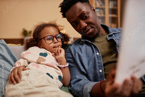 Medium shot of African American preschool girl in glasses on sofa at home interested in pictures of fairy tale book in fathers hands photo