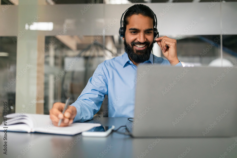 Cheerful indian man with headset working online on laptop - obrazy, fototapety, plakaty 