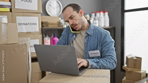 Hispanic bald man with beard using laptop and talking on phone in a donation center warehouse with boxes labeled 'donations'. photo