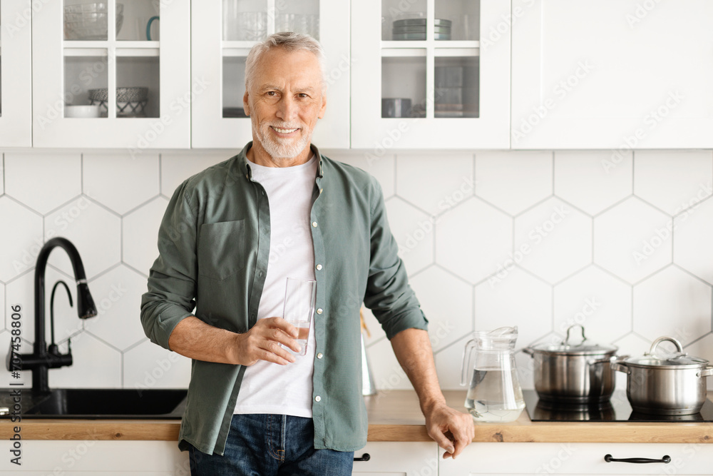 Cheerful mature man holding glass of water while posing in modern kitchen Cheerful mature man holding glass of water while posing in modern kitchen