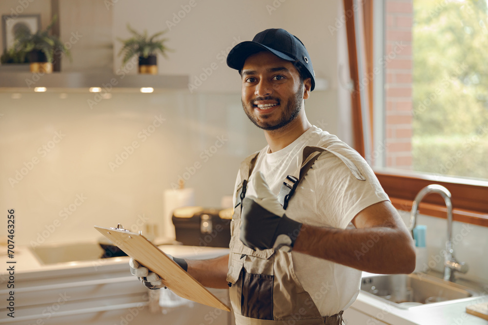 Portrait of young smiling repairman standing with clipboard and showing sign Ok in kitchen at home Portrait of young smiling repairman standing with clipboard and showing sign Ok in kitchen at home
