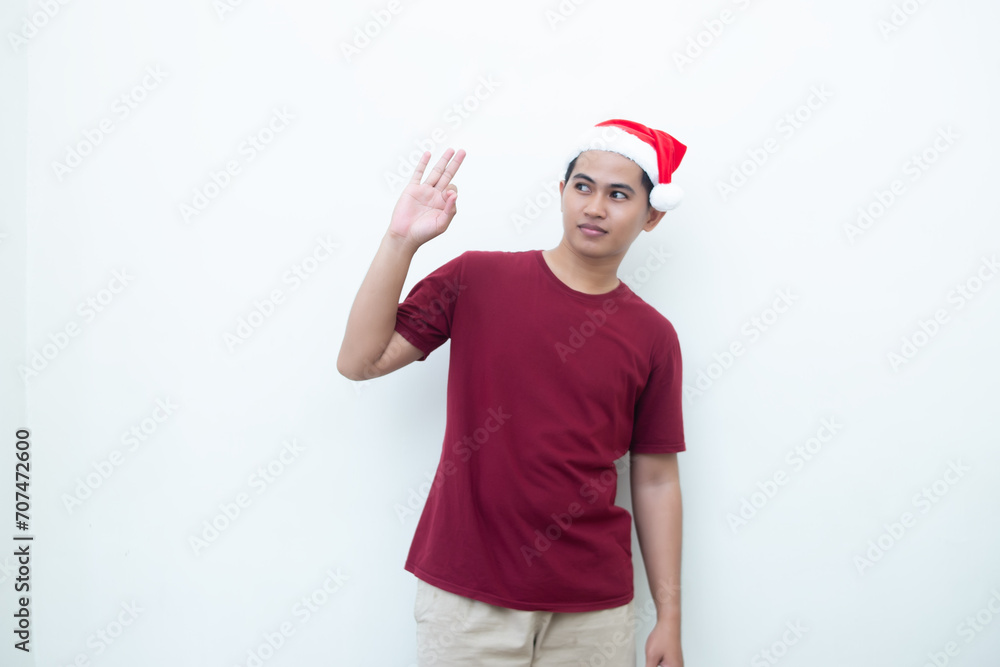 Young Asian man wearing a Santa Claus hat with a hand making the ok gesture isolated by a white background for visual communication Young Asian man wearing a Santa Claus hat with a hand making the ok gesture isolated by a white background for visual communication