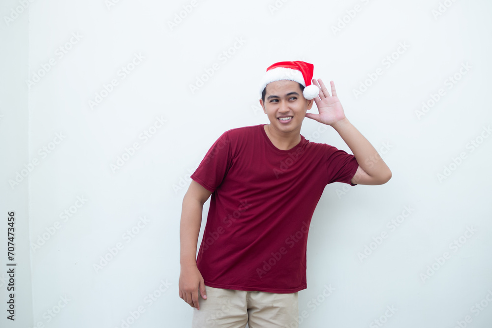 Young Asian man wearing a Santa Claus hat expressing listening with hand to ear isolated by a white background for visual communication
