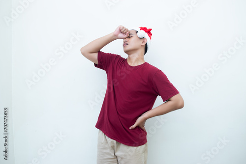 Young Asian man wearing a Santa Claus hat expressing a headache by holding his head and showing signs of pain isolated by a white background for visual communication