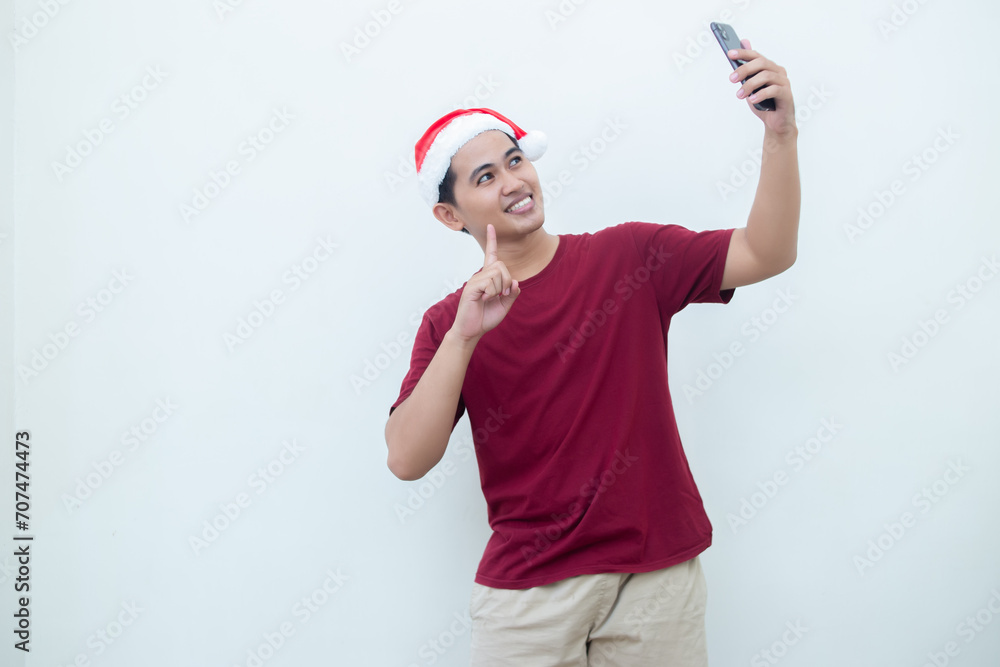 Young Asian man wearing a Santa Claus hat, holding a smartphone and taking a selfie isolated against a white background for visual communication Young Asian man wearing a Santa Claus hat, holding a smartphone and taking a selfie isolated against a white background for visual communication