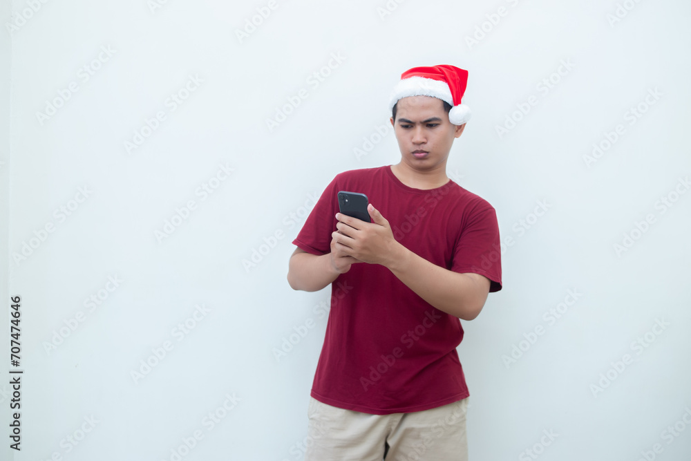 Young Asian man wearing a Santa Claus hat holding a smartphone and expressing a smile, shock and surprise isolated by a white background for visual communication