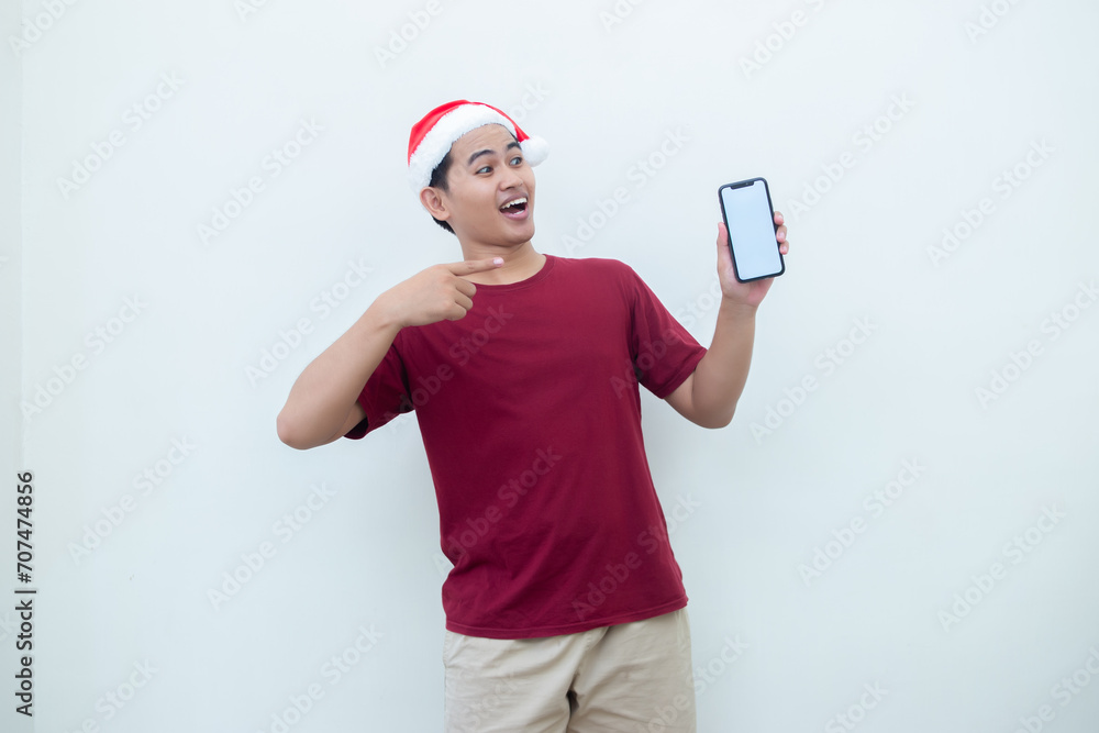 Young Asian man wearing a Santa Claus hat holding a smartphone, and pointing to the phone with a smile, look of shock, and surprise isolated against a white background for visual communication Young Asian man wearing a Santa Claus hat holding a smartphone, and pointing to the phone with a smile, look of shock, and surprise isolated against a white background for visual communication