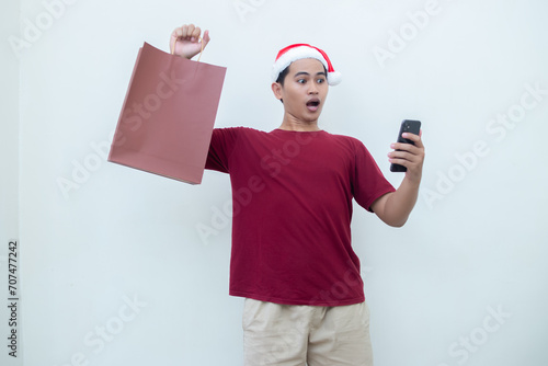 Young Asian man wearing a Santa Claus hat holding a smartphone and a shopping bag with expressions of smile, shock, and surprise, isolated against a white background for visual communication
