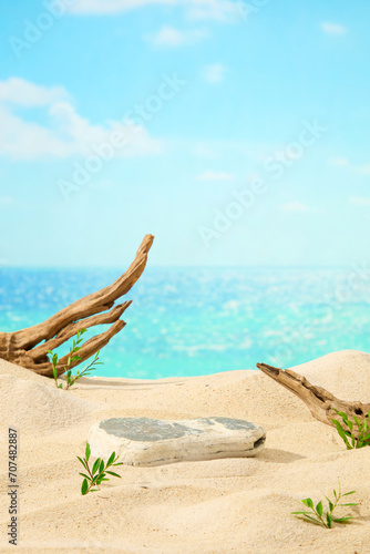 Front view of a gray block of stone displayed on sand beach background with dry twigs and green grass. Blue sea background and clear blue sky behind. Natural scene with space for display product