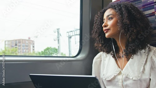 Young businesswoman commuting sitting on moving train passing through city working on laptop and listening to music or podcast on earphones looking out of window - shot in slow motion photo