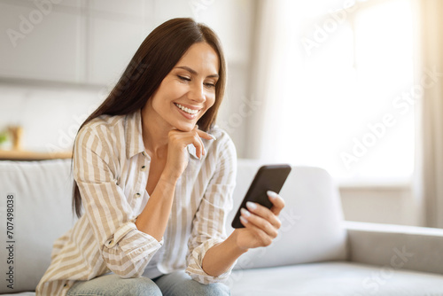 Smiling young woman using her smartphone while relaxing on couch at home