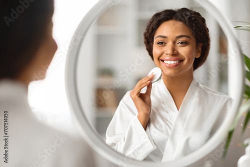 Beautiful black woman looking in mirror while cleansing skin with cotton pad photo