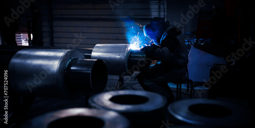 The welder works in the workshop with large-sized tube. Welding equipment and workwear. Sparks and red-hot metal particles flying around. photo