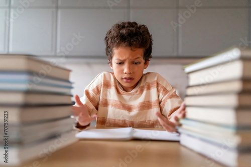 Puzzled black boy making homework surrounded by towering piles of books photo