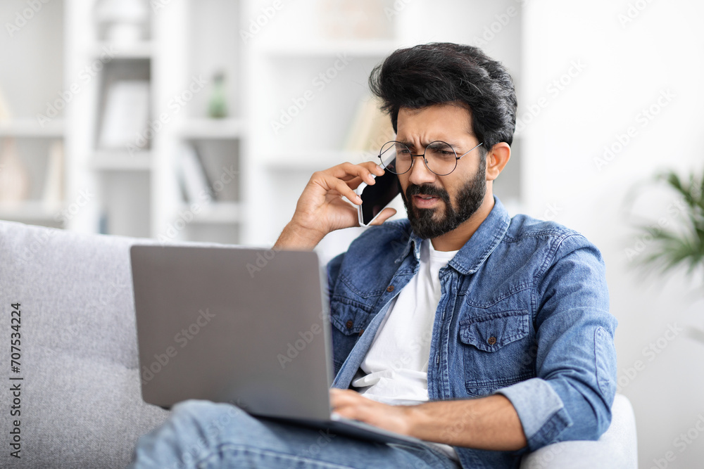 Annoyed indian man using phone while looking at his laptop at home Annoyed indian man using phone while looking at his laptop at home