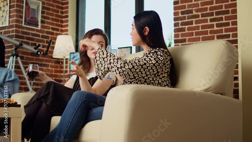 BFFs talking on couch, drinking alcohol beverages at friends social reunion and catching up with each other. Asian woman enjoying talking with mate, gossiping their boyfriends in modern living room photo