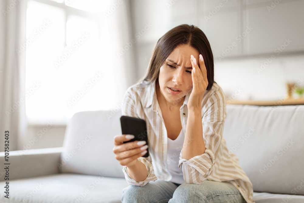 Worried young woman looking at smartphone screen at home and touching head Worried young woman looking at smartphone screen at home and touching head