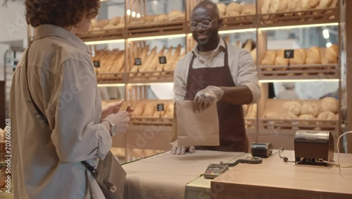 Medium shot of Caucasian female customer paying with card at bakery and Black salesman at counter smiling and giving her craft paper bag with takeout order photo