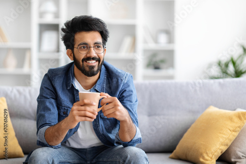 Joyful Indian man holding coffee cup with both hands and looking away