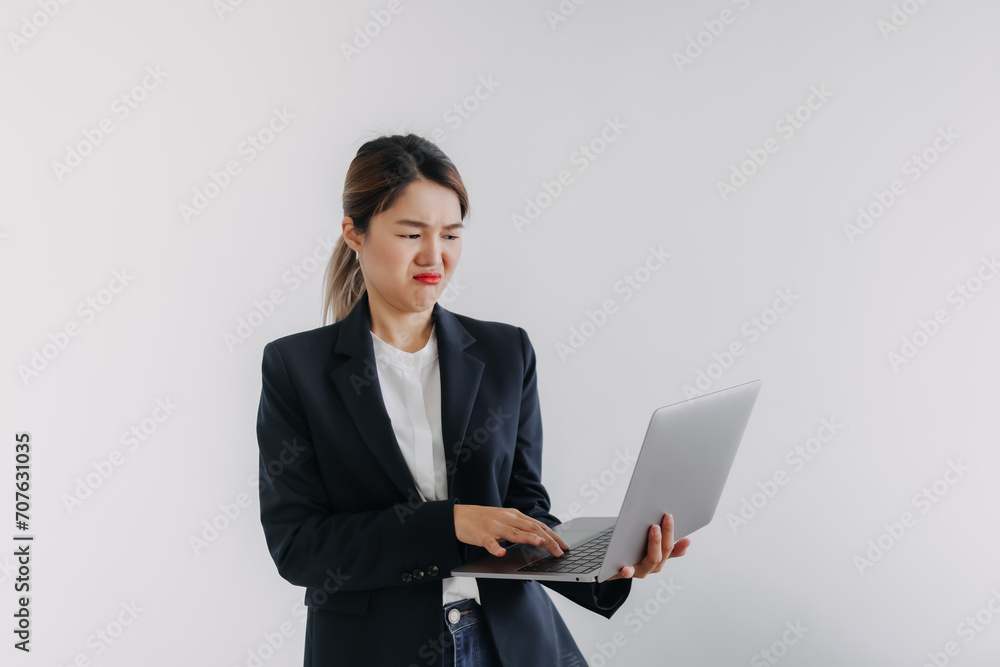 Unhappy asian Thai woman wear blazer suit, looking above at laptop with sad bored face, dislike holding computer, standing over white background wall. Unhappy asian Thai woman wear blazer suit, looking above at laptop with sad bored face, dislike holding computer, standing over white background wall.