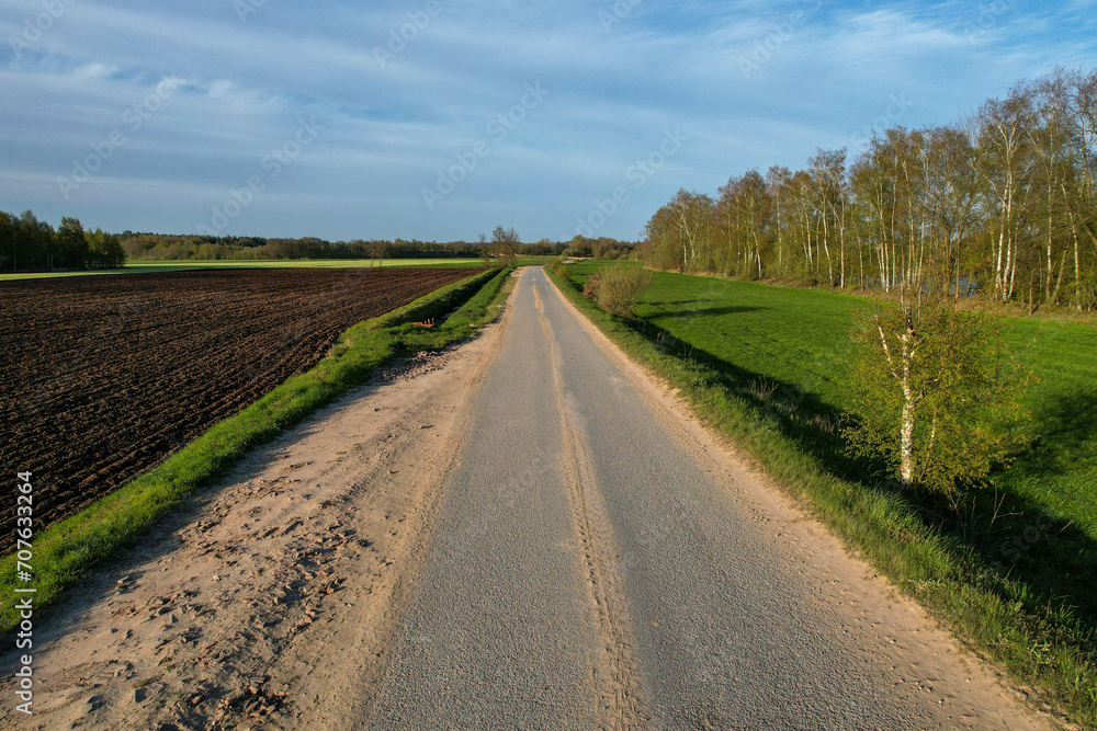 This image presents a straight country road dividing contrasting landscapes: on one side, there is a plowed field, ready for planting, indicative of the agricultural activity that defines the area; on This image presents a straight country road dividing contrasting landscapes: on one side, there is a plowed field, ready for planting, indicative of the agricultural activity that defines the area; on