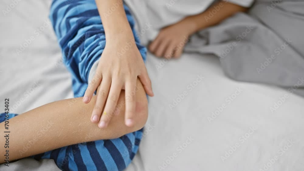 Adorable blond kid touching injured knee, upset and suffering in pain, lying on bedroom bed, worry etched on his little face Adorable blond kid touching injured knee, upset and suffering in pain, lying on bedroom bed, worry etched on his little face