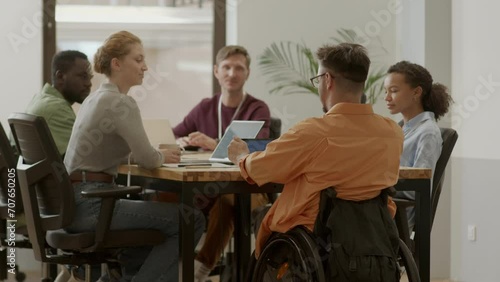 Male team leader in wheelchair talking to group of multi-ethnic colleagues while holding business meeting in office photo