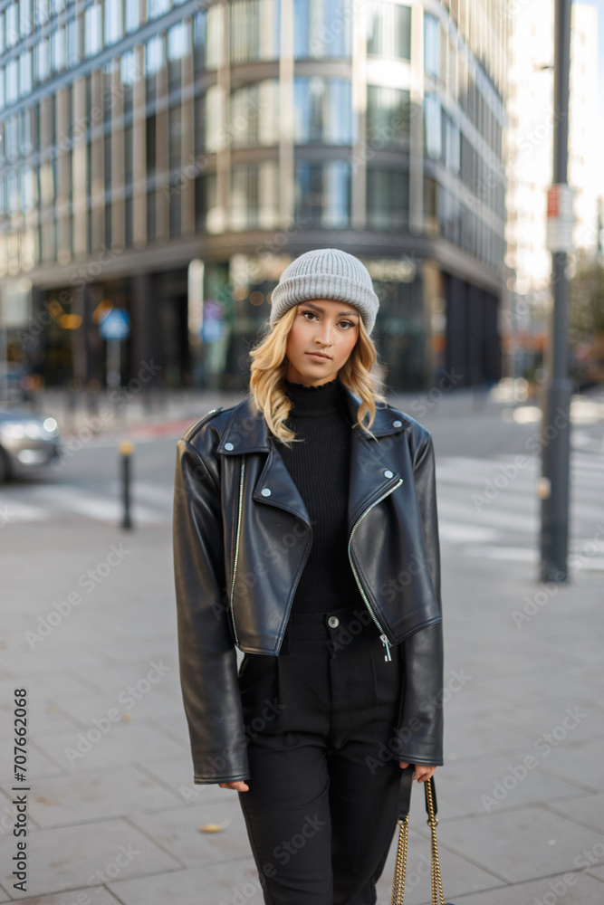 Fashion beautiful urban stylish girl model with a knitted hat in a black knitted sweater and leather jacket walks in the city