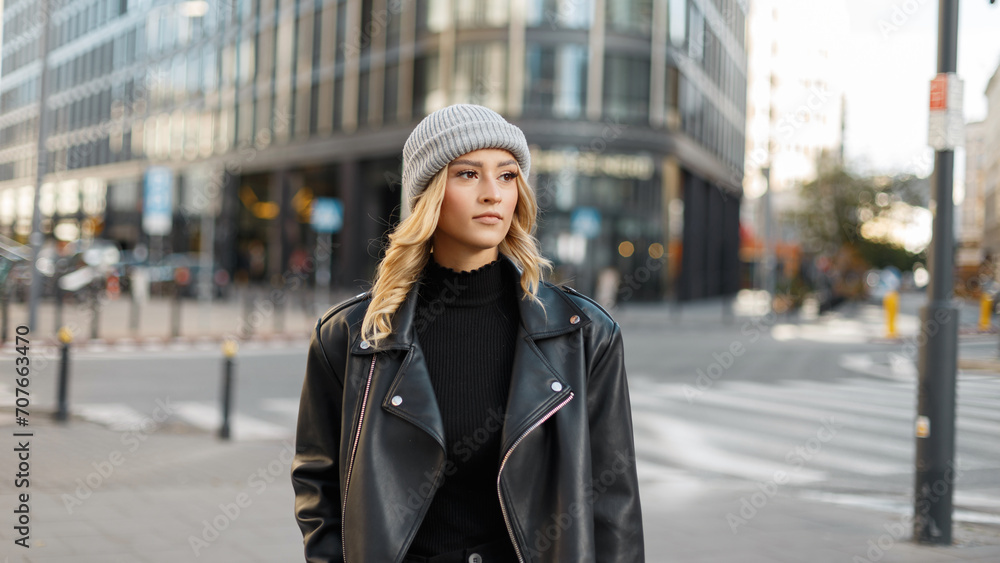 Beautiful fashionable girl model in a stylish knitted hat in a black leather fashion jacket walks in the city