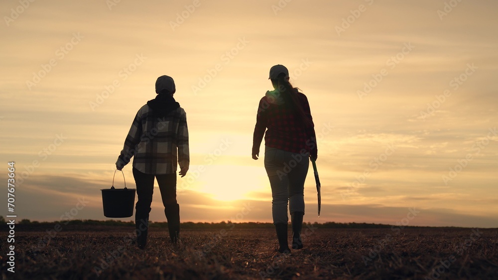 Owners farm with shovel and bucket go through field. Agronomic work on plantation. Agriculture. Two farmers in boots walking across field. Teamwork in field at sunset. Concept of agricultural business